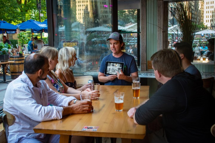Group of people at a wooden table, one holding a tablet, surrounded by drinks in a pub setting.